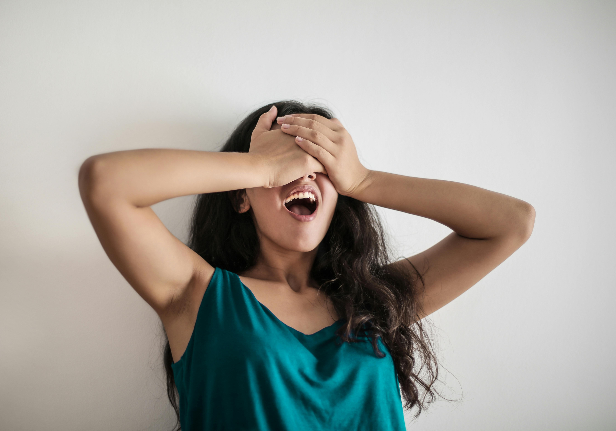 Woman in teal shirt covering her face with her hands on a plain background