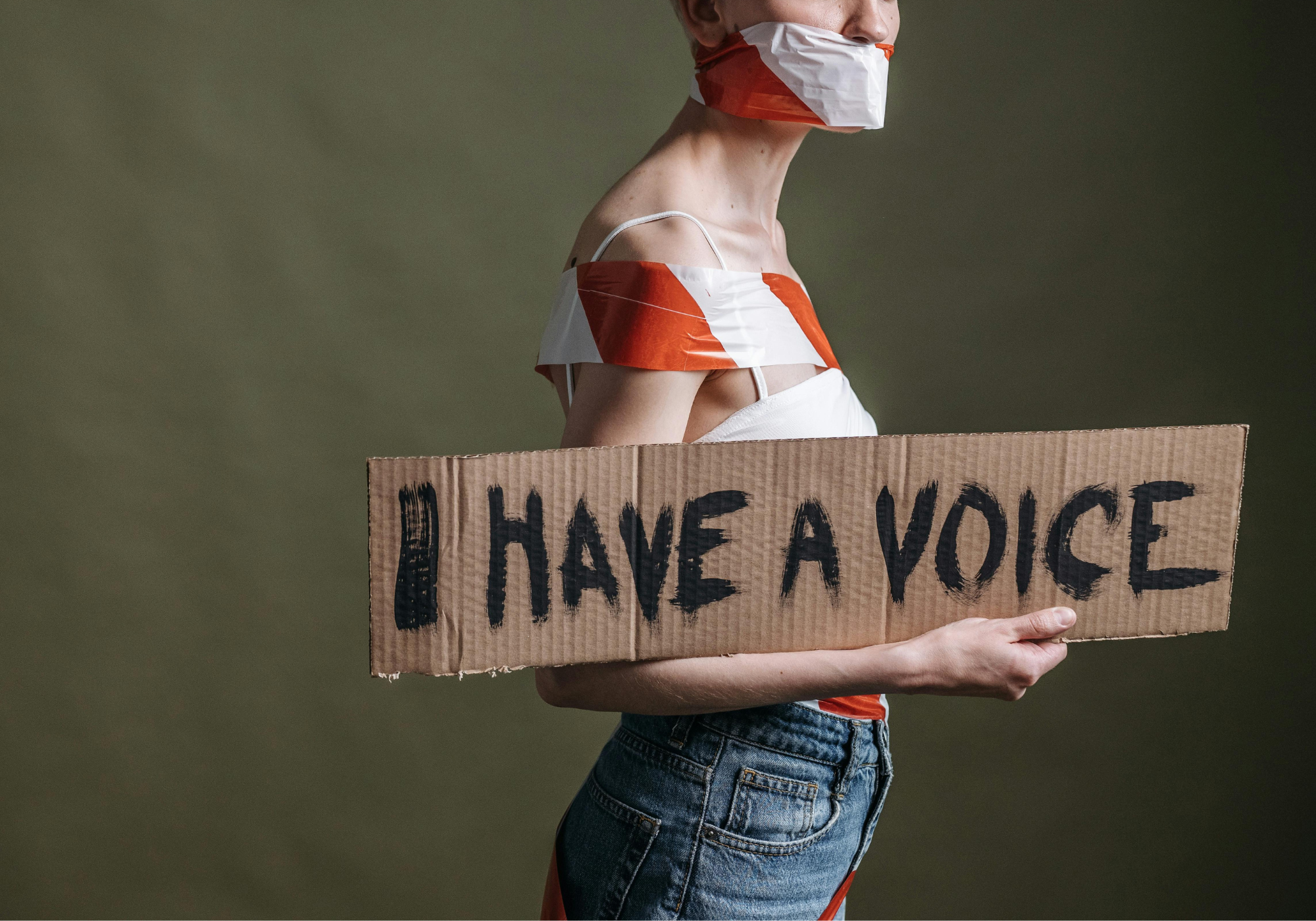 Person wearing a face mask and holding a cardboard sign with 'I have a voice' text against a plain background