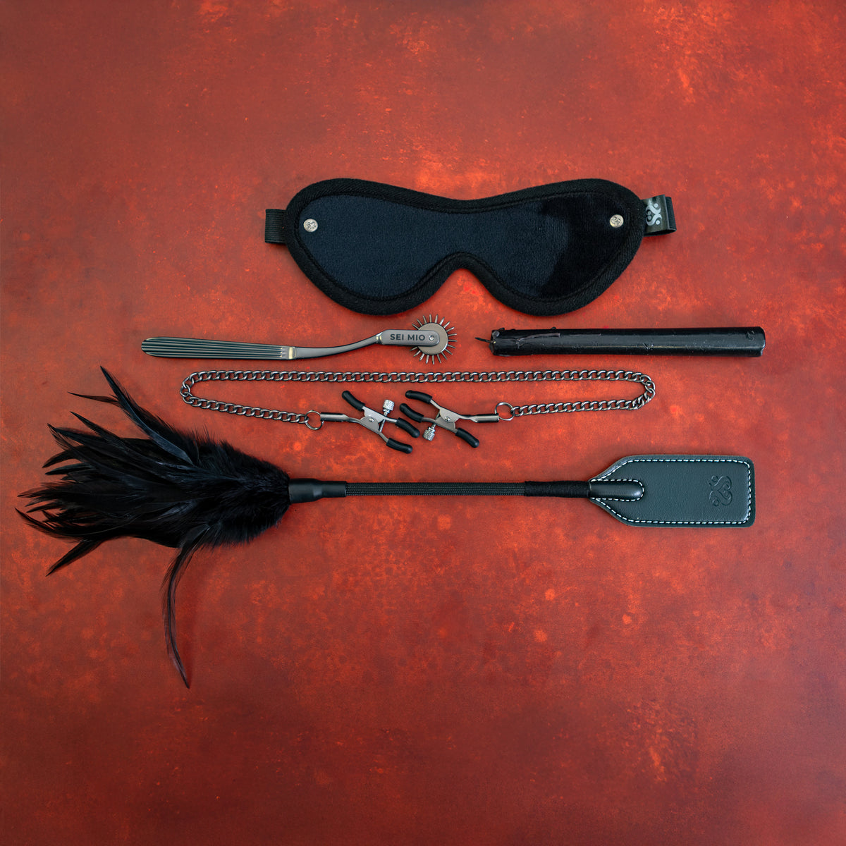 Sei Mio Fetish Feels kit on red: blindfold, pinwheel, candle, chained nipple clamps and feather crop tickler