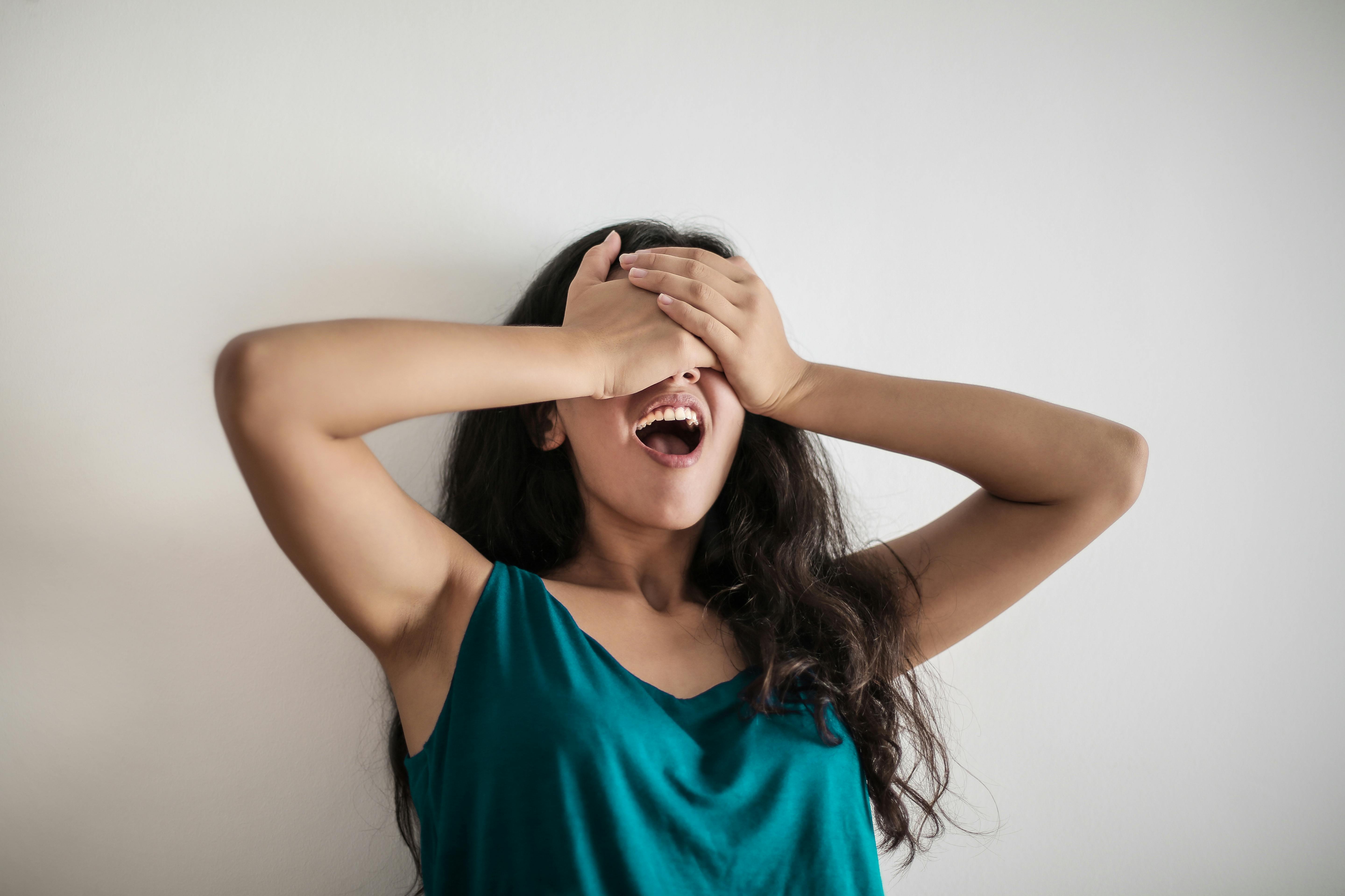 Woman in teal shirt covering her face with her hands against a plain background