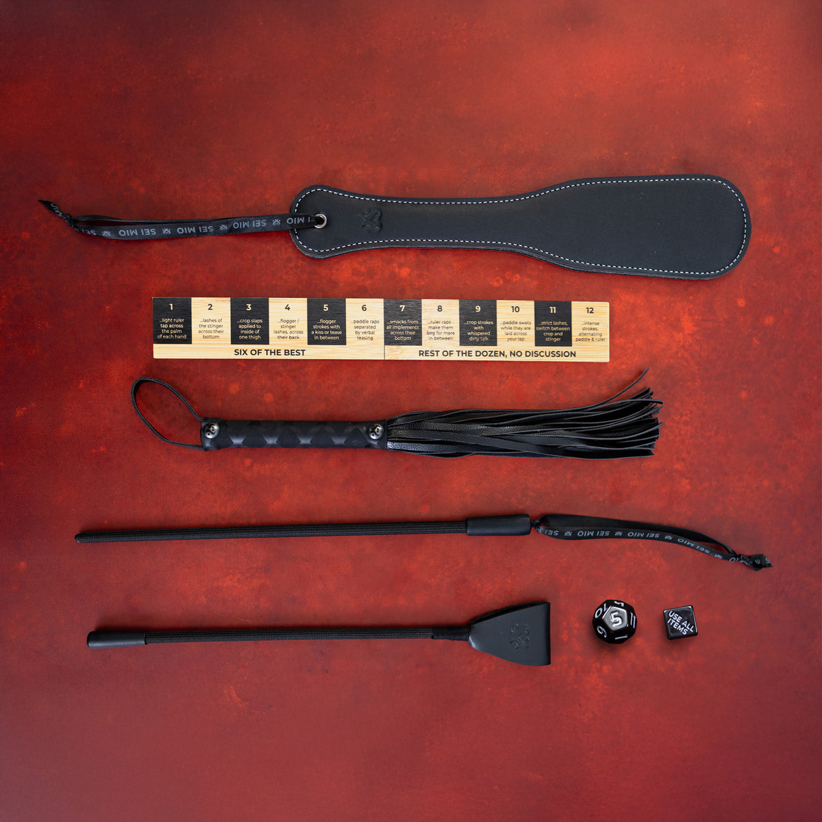 Sei Mio Correction Collection on red: spanking paddle, bamboo ruler, flogger, stinger, crop and two dice