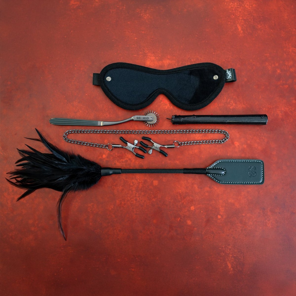 Sei Mio Fetish Feels kit on red: blindfold, pinwheel, candle, chained nipple clamps and feather crop tickler